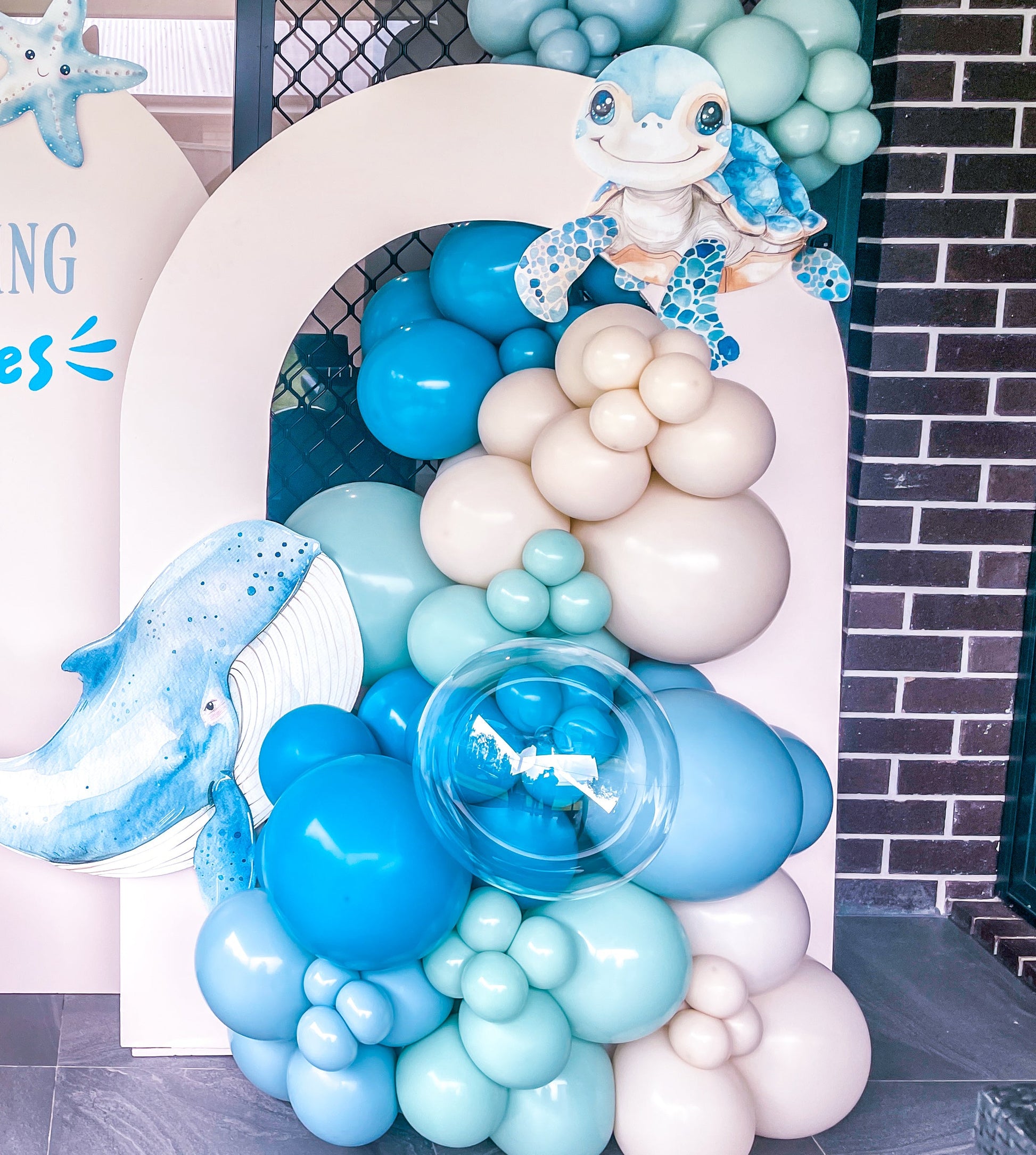 Balloon arch with blue, white, and beige balloons and ocean-themed decorations in front of a building.
