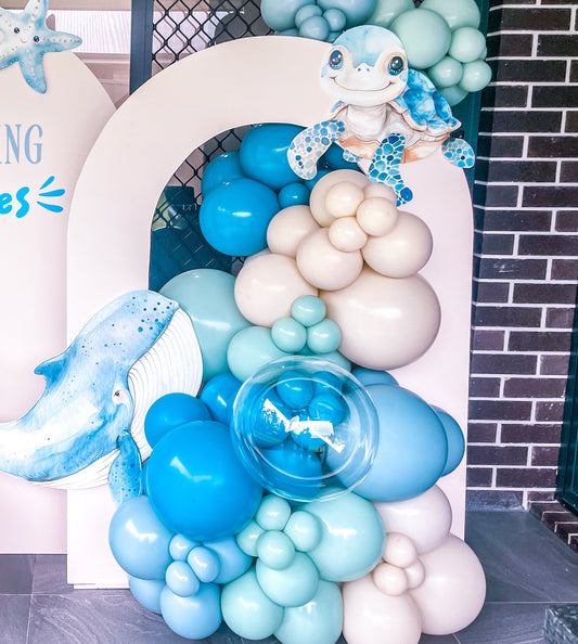 Balloon arch with blue, white, and beige balloons and ocean-themed decorations in front of a building.